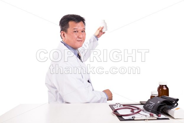 Matured Asian medical practitioner holding medicine seated behind desk - ThamKC Royalty-Free Photos